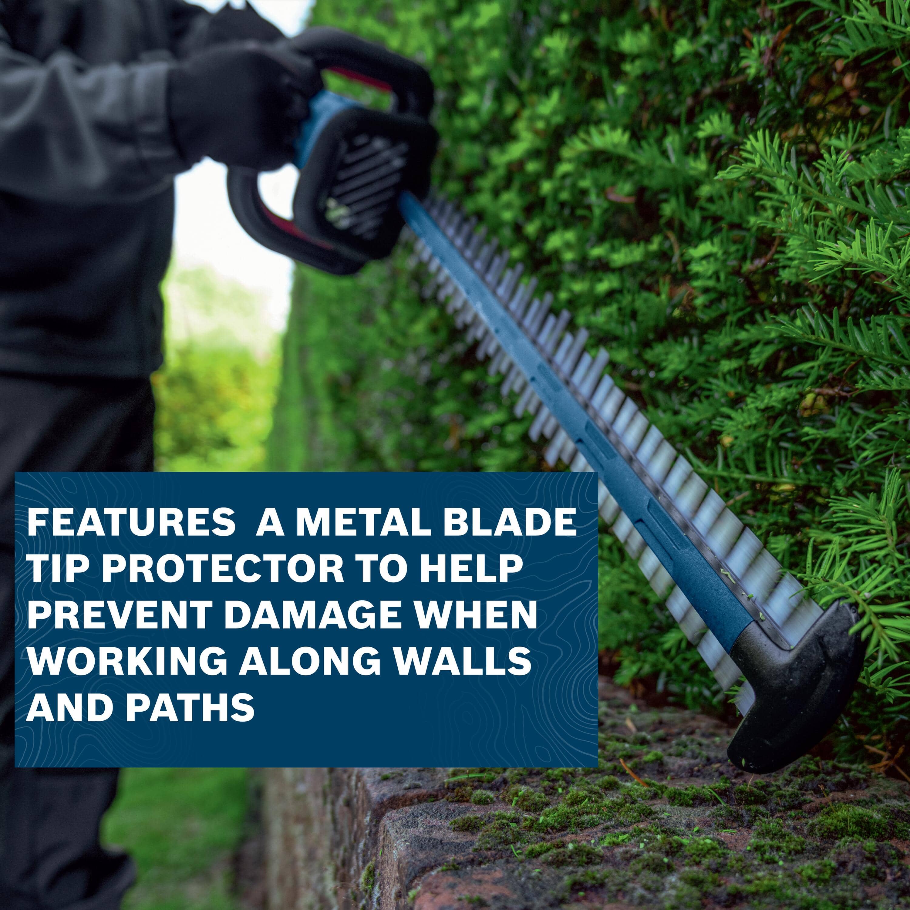 An operational shot demonstrating close-clearance landscaping trimming along a perimeter border, showing the trimmer blade tracking parallel to a moss-covered structural brick base wall beneath a dark blue banner reading FEATURES A METAL BLADE TIP PROTECTOR TO HELP PREVENT DAMAGE WHEN WORKING ALONG WALLS AND PATHS.