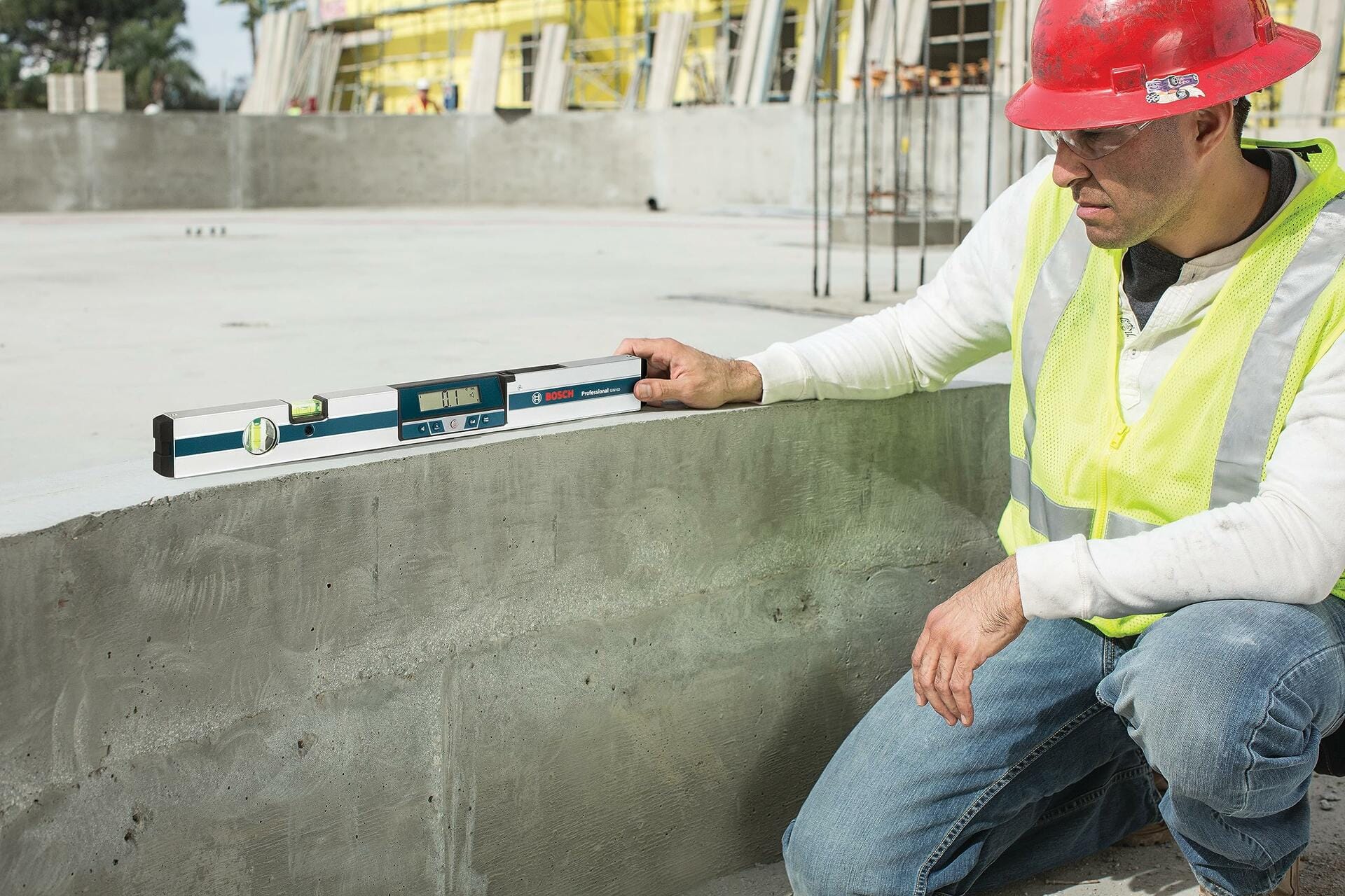 Worker in safety vest and hard hat using the Bosch GIM60 digital level to verify the horizontal plane of a newly poured concrete foundation on an active jobsite