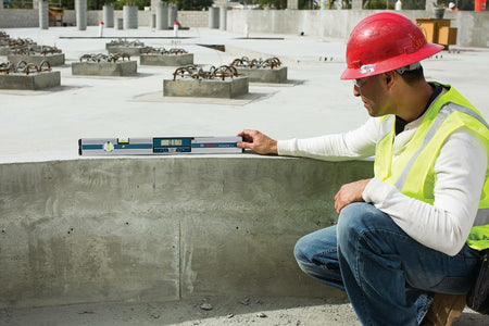 Construction worker kneeling on a jobsite and using the Bosch GIM60 digital level to check the horizontal alignment and level of a large finished concrete slab or foundation wall