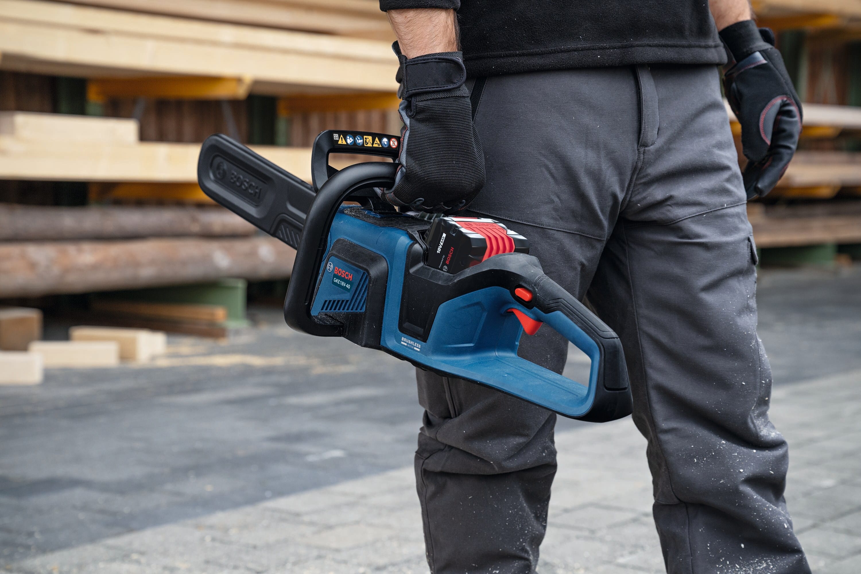 A close-up view of a professional property manager wearing grey work trousers and protective gloves holding the compact Bosch 18V cordless chainsaw by its top handle handle at his side, showcasing its portable lightweight design and clean battery compartment layout.