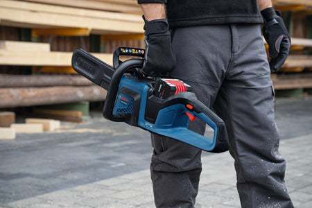 A close-up view of a professional property manager wearing grey work trousers and protective gloves holding the compact Bosch 18V cordless chainsaw by its top handle handle at his side, showcasing its portable lightweight design and clean battery compartment layout.