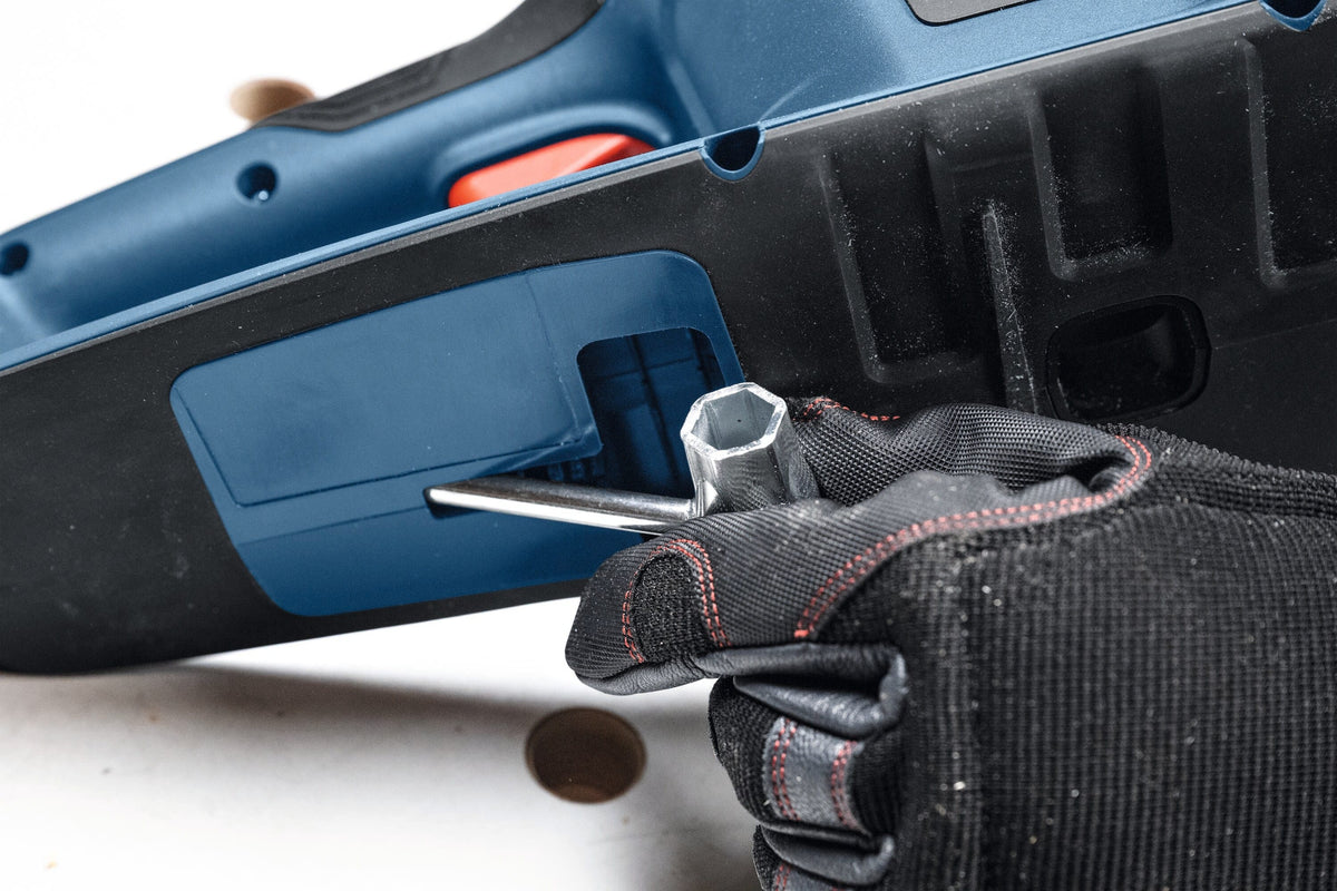 A detailed macro close-up view highlighting the underside of the blue chainsaw chassis as a gloved hand slides the dual-ended steel scrench service tool into its dedicated onboard storage slot pocket.