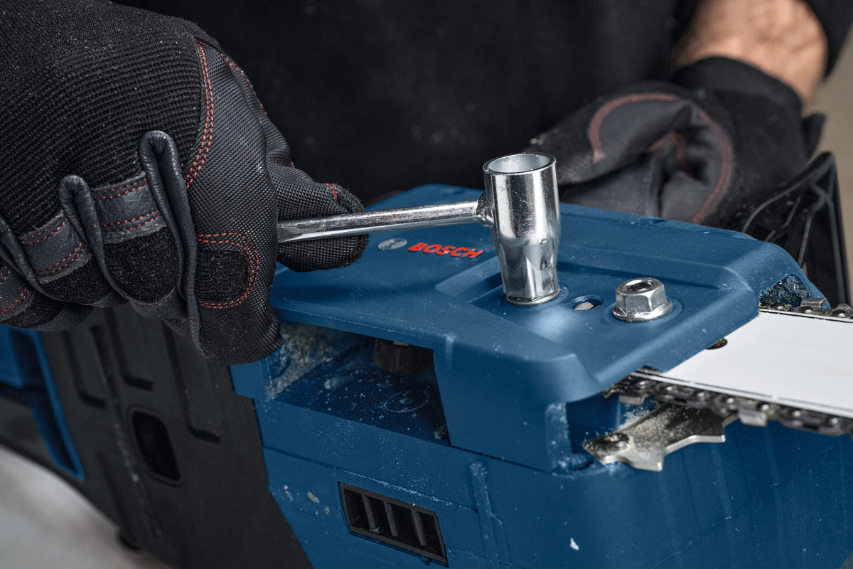 A close-up maintenance shot showing a woodcutter using the metal socket scrench key tool to tighten down the locking hex nut on the side sprocket tensioning cover plate of the blue Bosch chainsaw body.