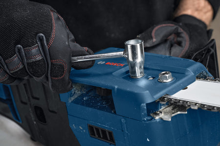 A close-up maintenance shot showing a woodcutter using the metal socket scrench key tool to tighten down the locking hex nut on the side sprocket tensioning cover plate of the blue Bosch chainsaw body.