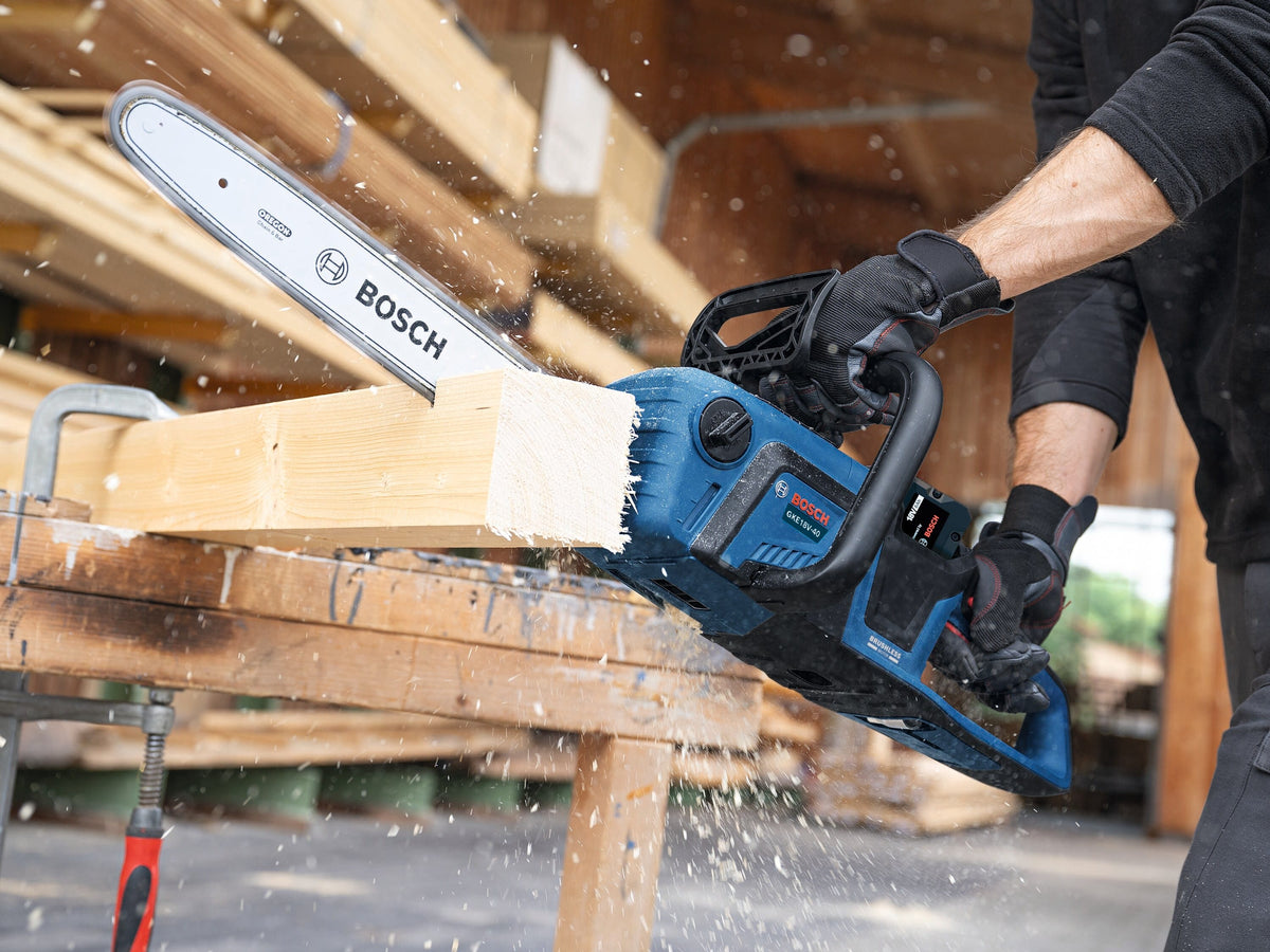 A high-speed action view of the Bosch brushless 18V chainsaw ripping cleanly through a thick, square architectural timber beam clamped to a jobsite workbench table, capturing a dramatic spray of flying wood shavings and saw dust.