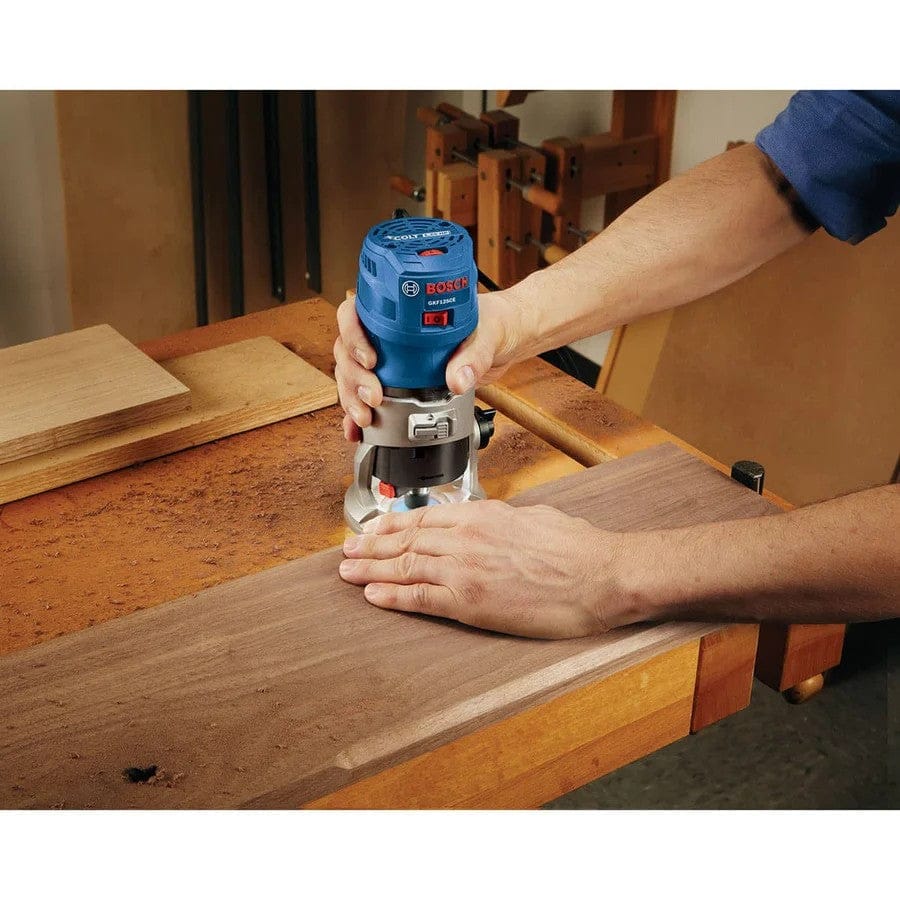 Person using Bosch palm router on wood clamped to workbench; wood shavings and clamps visible in workshop setting.