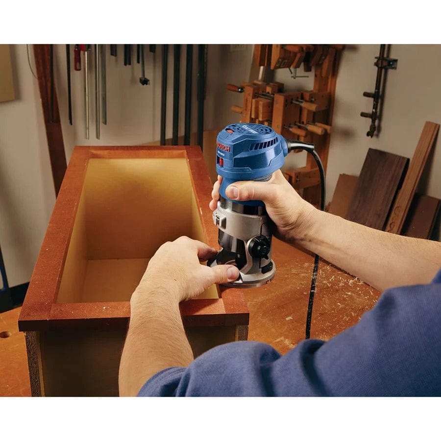 Person shaping edge of wooden cabinet with Bosch router; workshop wall features clamps and woodworking tools.