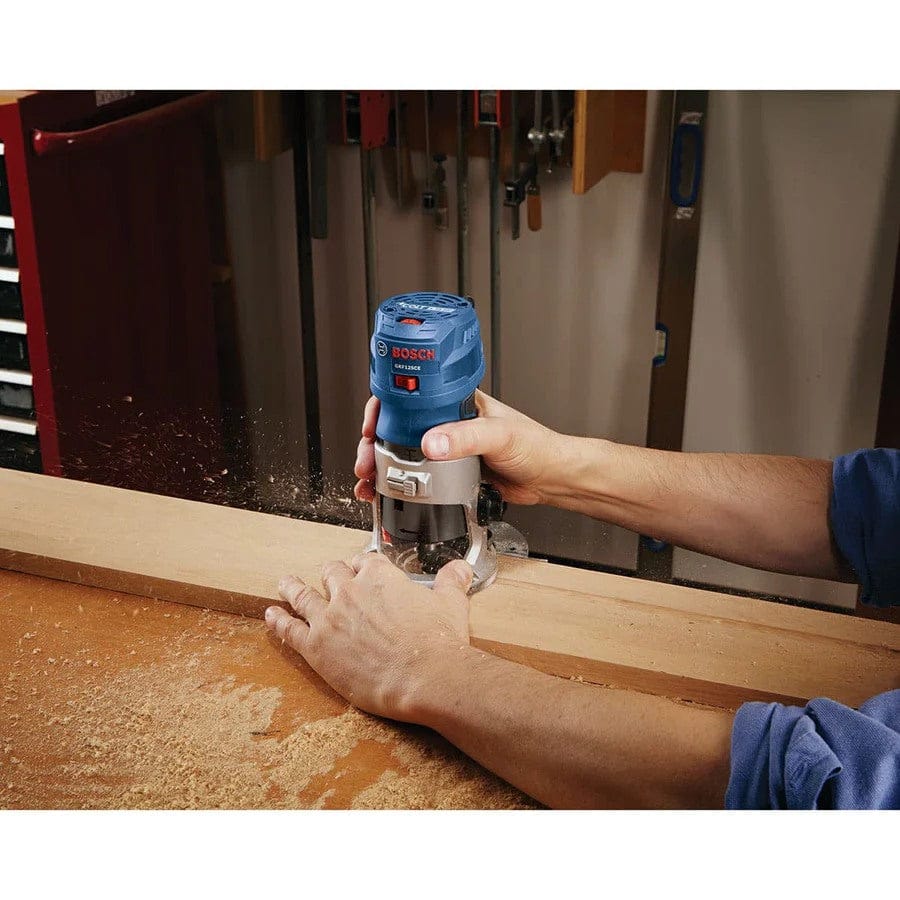 Individual guiding Bosch palm router along wooden board edge; wood dust and hanging tools visible in background.