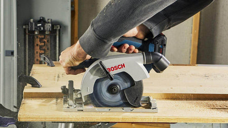 An action shot of a person using the Bosch circular saw to cut a piece of plywood on a jobsite, showing the blade-left design providing a clear view of the cut line for the right-handed operator.