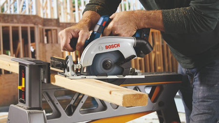 An operator using the circular saw to cut a 2x4 piece of lumber supported on a sawhorse, illustrating the 2-1/4 inch cut capacity of the 6-1/2 inch blade.