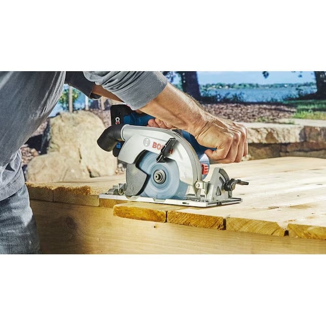 Person using Bosch circular saw to cut thick wooden plank outdoors; stone wall and trees visible in background.