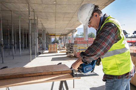 An outdoor application lifestyle photo of a commercial construction builder dressed in a white safety hard hat, protective glasses, a flannel shirt, and a high-visibility yellow vest, using the Bosch 18V cordless circular saw to execute a straight crosscut on a structural lumber plank resting over saw horses.