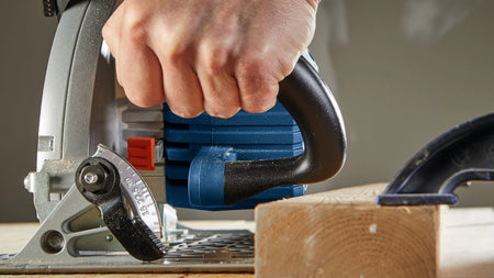 A close-up detail shot focusing on a contractor's hand holding the thick black front auxiliary handle of the blue Bosch saw, demonstrating the tight physical control grip positioned adjacent to the front quick-locking bevel level adjustment assembly.