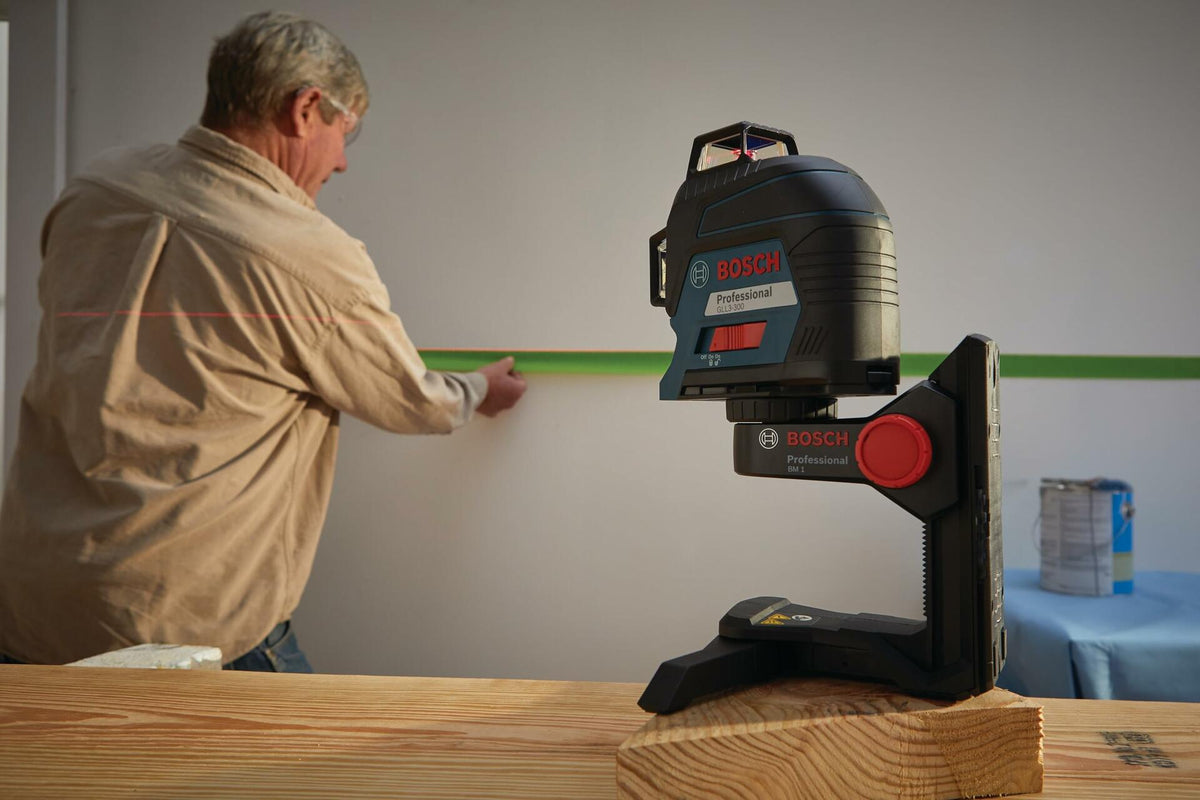 Worker applying green tape along a red laser line projected by a Bosch laser level mounted on a stand. The scene includes painting supplies, emphasizing the tool’s use in layout and alignment for renovation.