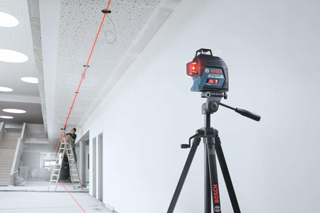 Bosch laser level mounted on a tripod projecting a red laser line across a ceiling and floor in a modern construction setting. A worker on a ladder aligns fixtures along the beam, demonstrating full-room layout capabilities.
