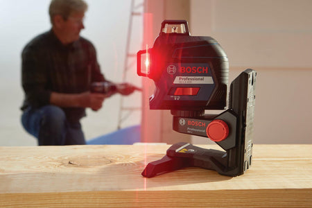 Bosch GLL 3-80 laser level emitting a red horizontal beam across a construction space. A person in the background uses a power drill, highlighting the tool’s role in precise alignment during active work.