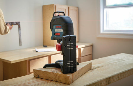 Bosch laser level placed on a wooden surface projecting a red horizontal line across a room. A person uses a square tool to align cabinetry, showcasing the tool’s precision in interior renovation.