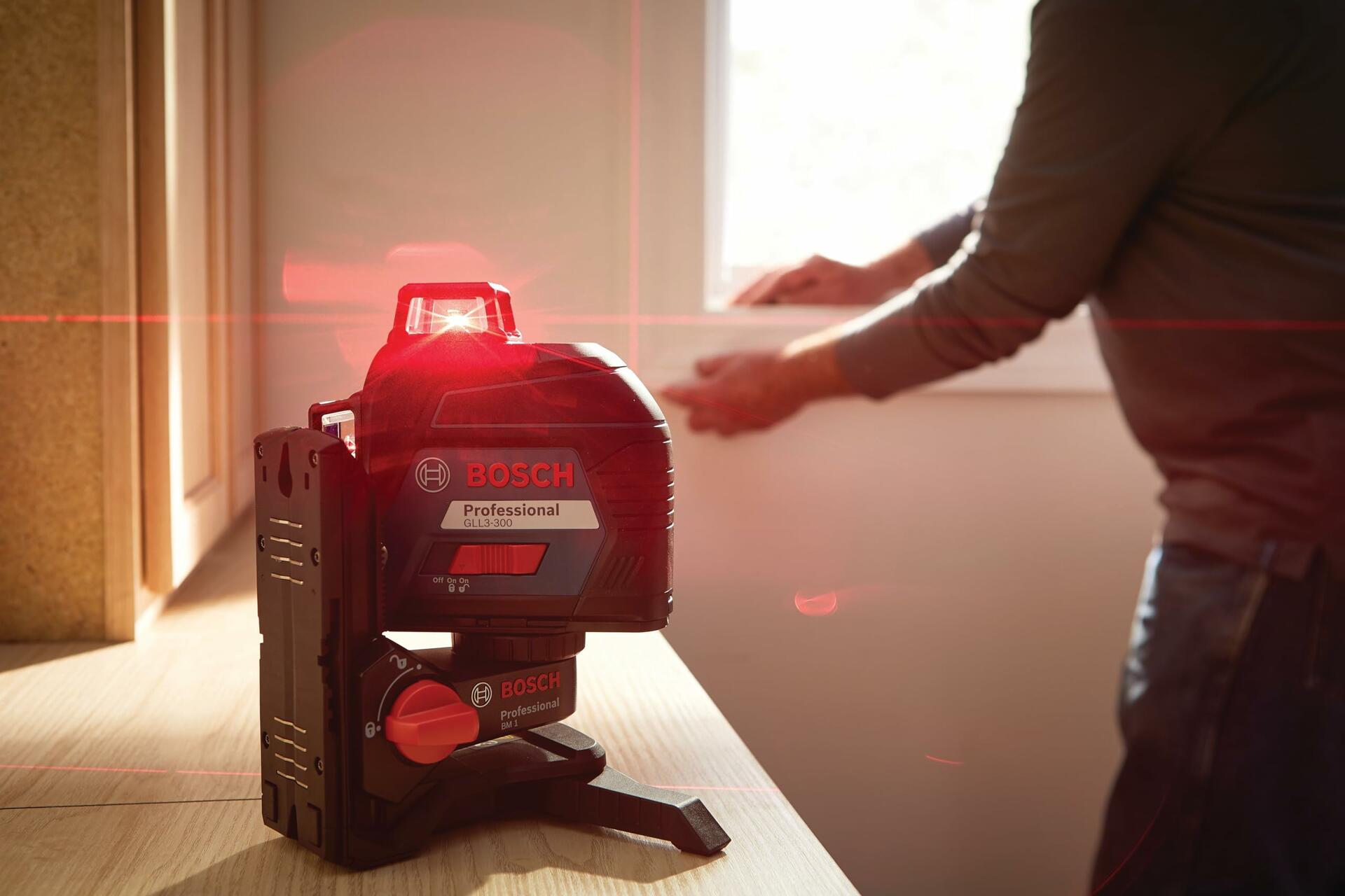 Bosch laser level projecting intersecting red horizontal and vertical lines. A worker in the background uses the alignment for installation tasks. The device is mounted for stability on a wooden surface.