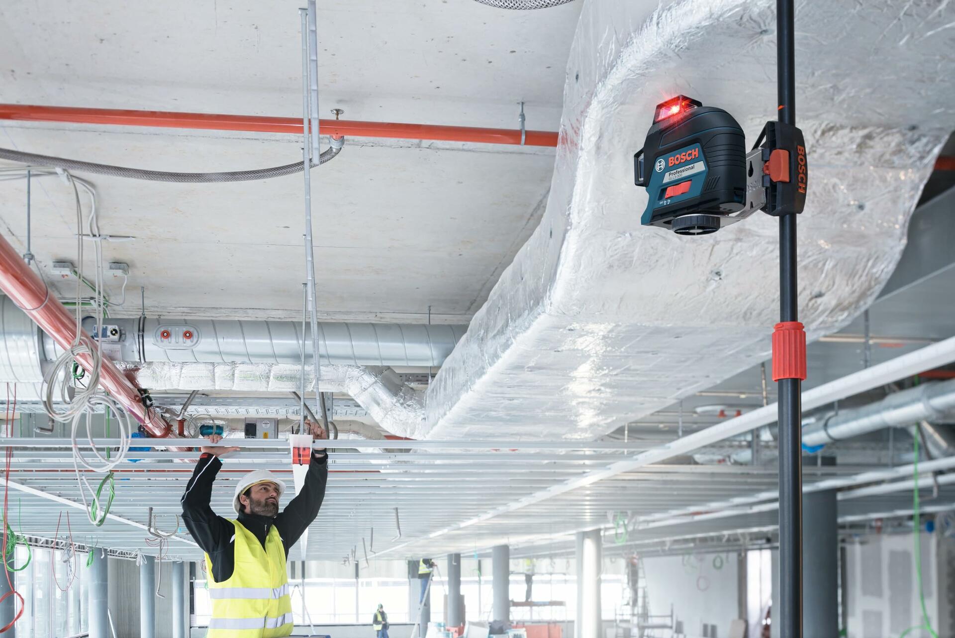 Construction worker in safety gear adjusting ceiling fixtures while a Bosch laser level projects a red alignment line. The device is mounted on a vertical pole, aiding precise installation of pipes and conduits.