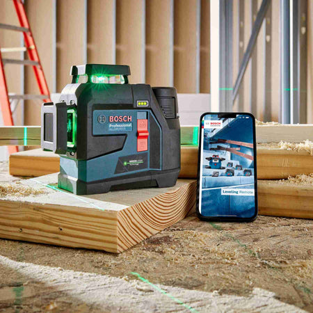 A close-up view of the laser level sitting on wooden framing lumber at a construction site, projecting bright green vertical and horizontal lines, positioned next to a smartphone displaying the connected leveling remote interface.