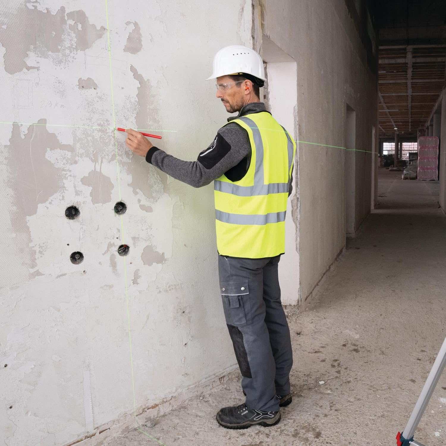 A construction worker in a safety vest marking a wall along the green laser beam projected by the tool