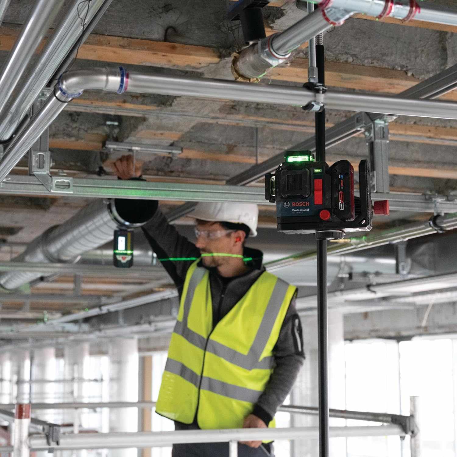 The laser level mounted high on a telescoping pole during a ceiling installation with a worker checking alignment in the background