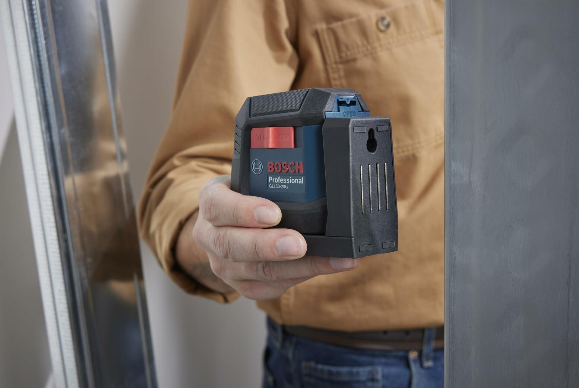 Person holding Bosch GLL 2-15 G laser level in a construction setting with metal framing, demonstrating tool use for alignment tasks.