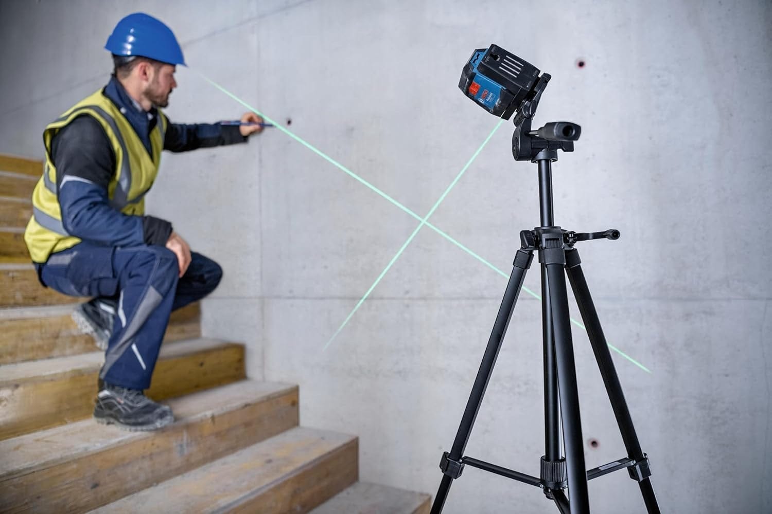 Jobsite scene showing a worker on a staircase using the laser level mounted on a tripod to project diagonal green reference lines on a concrete wall.