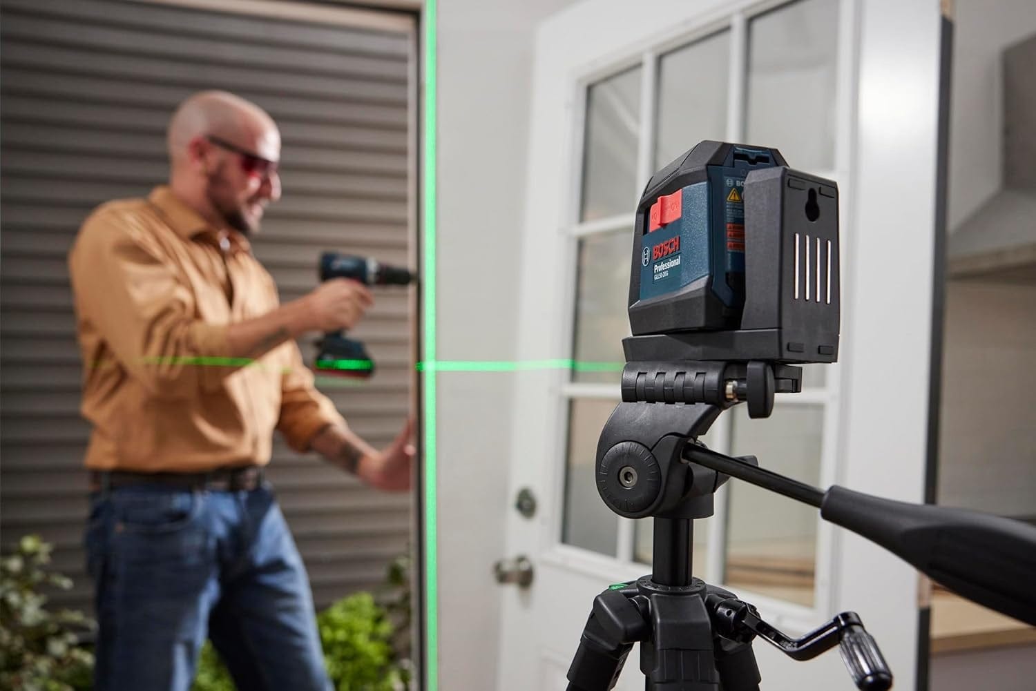 Bosch GLL3-330CG laser level mounted on tripod projecting green horizontal line; person in background aligns work near door frame.