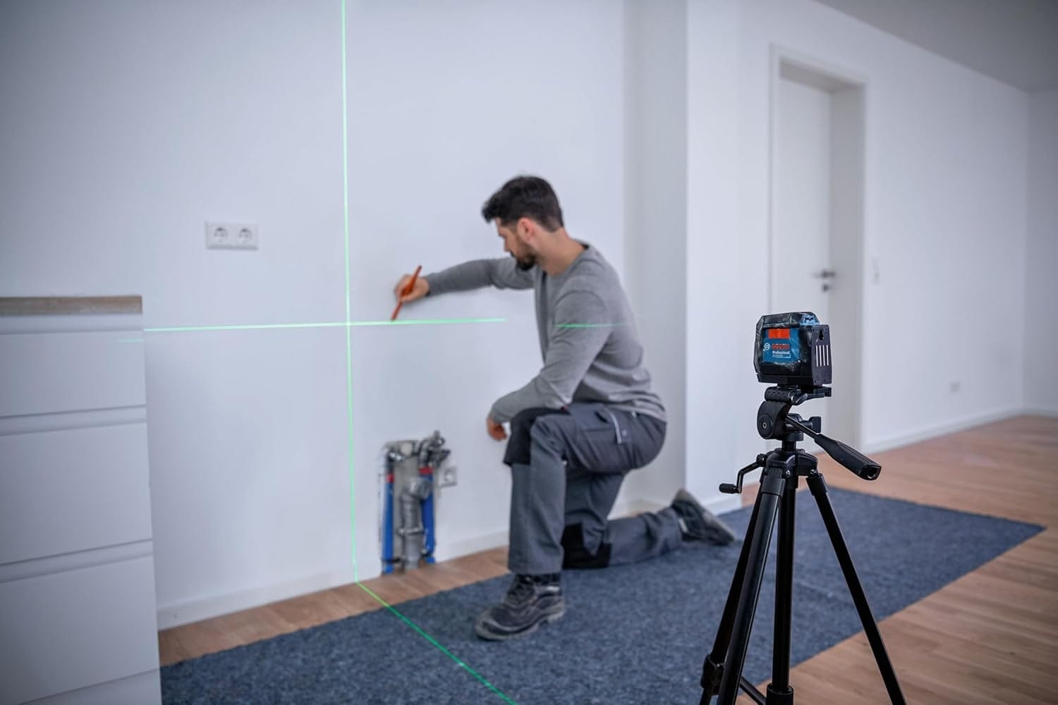 Person marking a white wall using a Bosch laser level mounted on a tripod; green cross-line beams guide alignment near exposed plumbing.