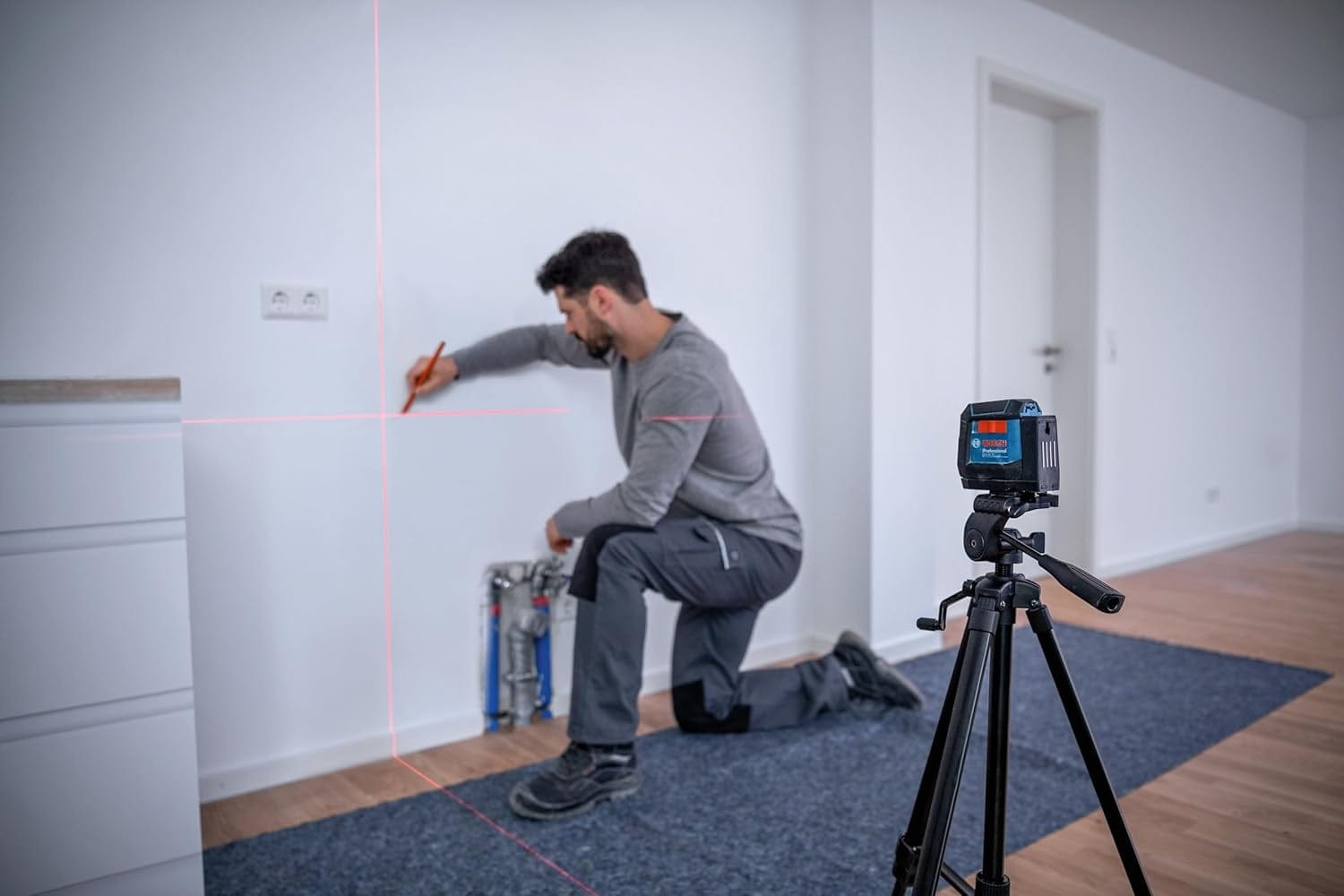 A construction professional kneeling in a room using the red cross-line beams from a tripod-mounted Bosch GLL50-20 laser level to mark precise plumbing or electrical points on a wall.
