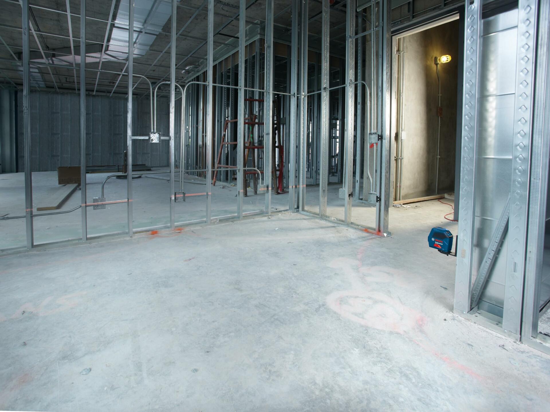 The Bosch GLL 55 self-leveling laser level magnetically attached to a vertical steel stud in a large commercial jobsite, projecting a steady red horizontal leveling line across the room.