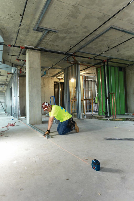A contractor on a jobsite utilizing the Bosch GLL 55 laser level on the floor to align metal track framing with a visible red laser line for accurate interior wall layout.