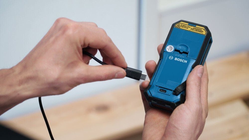 Close-up of a person's hands connecting a USB-C charging cable into the charging port of a Bosch GBA37V10 Lithium-Ion battery pack for BLAZE laser measures.