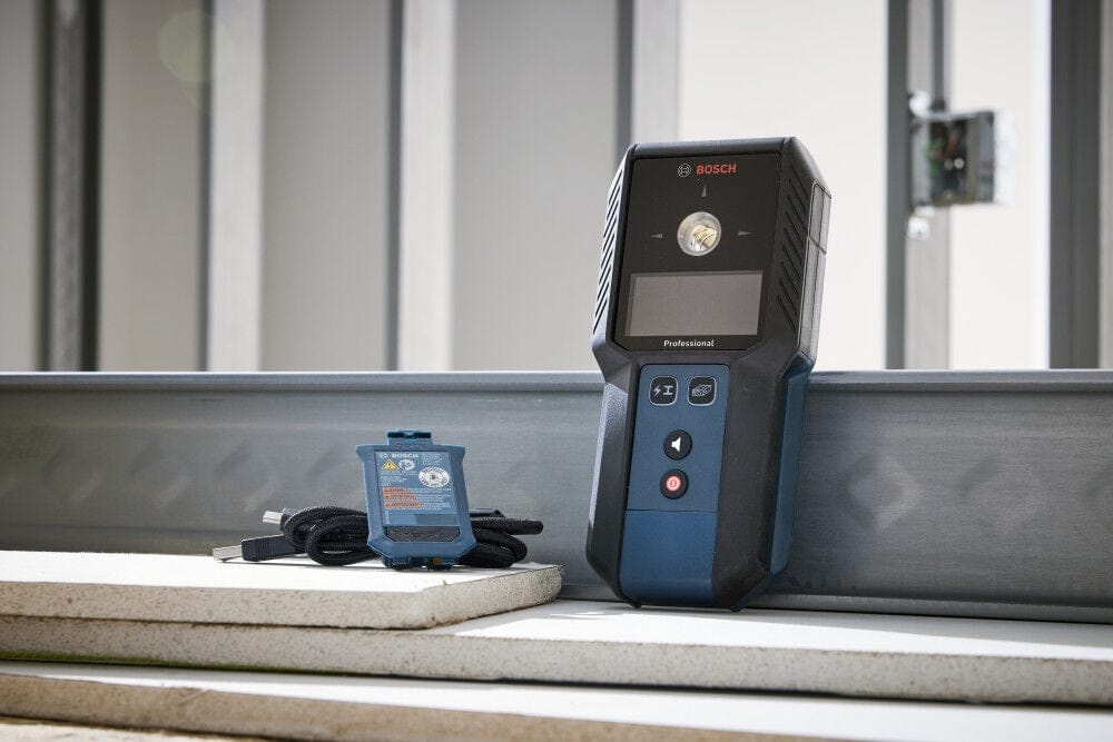 A Bosch Professional laser measure leaning against a metal stud on a construction site next to a Bosch GBA37V10 rechargeable battery and a coiled USB-C charging cable.