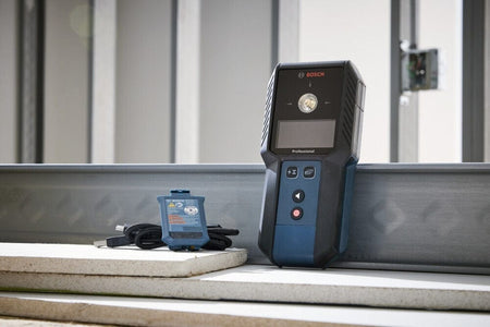 A Bosch Professional laser measure leaning against a metal stud on a construction site next to a Bosch GBA37V10 rechargeable battery and a coiled USB-C charging cable.