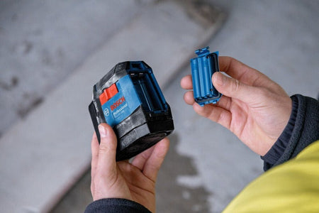 A person holding a Bosch Professional GLL50-20G line laser in one hand and a blue Bosch GLM-BAT 3.7V Lithium-Ion battery pack in the other, demonstrating how the battery replaces standard AA batteries.