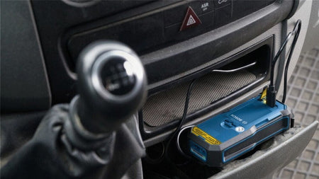 A Bosch BLAZE laser measure powered by a GLM-BAT lithium-ion battery pack charging via a USB cable while resting in a vehicle's center console storage compartment.