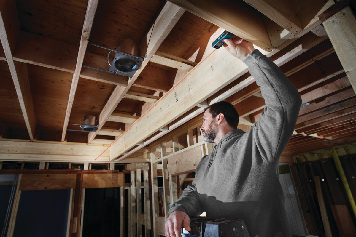 An on-site residential construction application photograph of a framing carpenter standing on a step stool and aiming the handheld Bosch GLM 20 laser distance tool upward against a wood ceiling joist to map out an unlit overhead electrical rough-in installation.