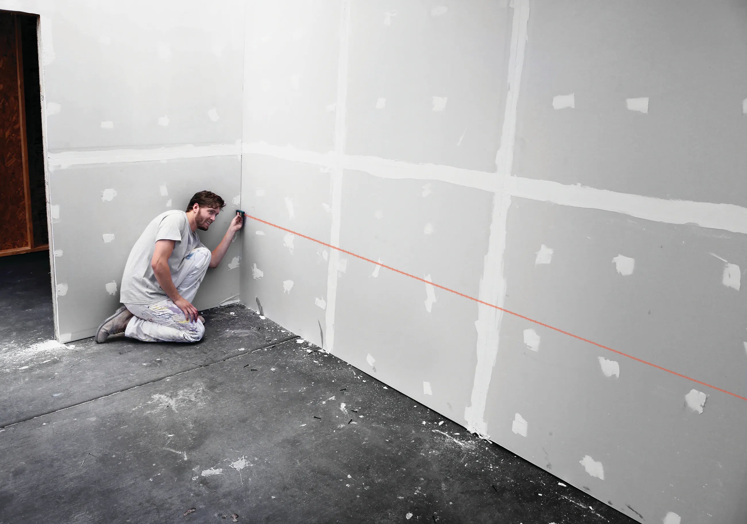 A real-world interior application photograph showing a drywall installer kneeling on a raw concrete floor deck and holding the blue Bosch digital laser measure flush against a corner stud to project a straight red tracking baseline across finished drywall panels.