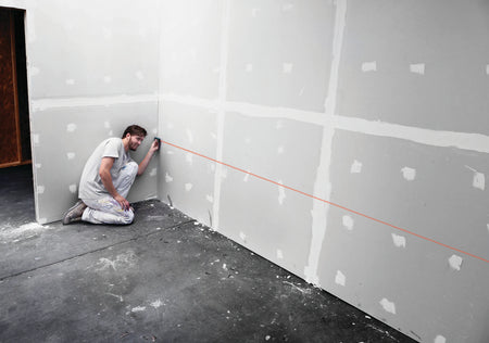 A real-world interior application photograph showing a drywall installer kneeling on a raw concrete floor deck and holding the blue Bosch digital laser measure flush against a corner stud to project a straight red tracking baseline across finished drywall panels.