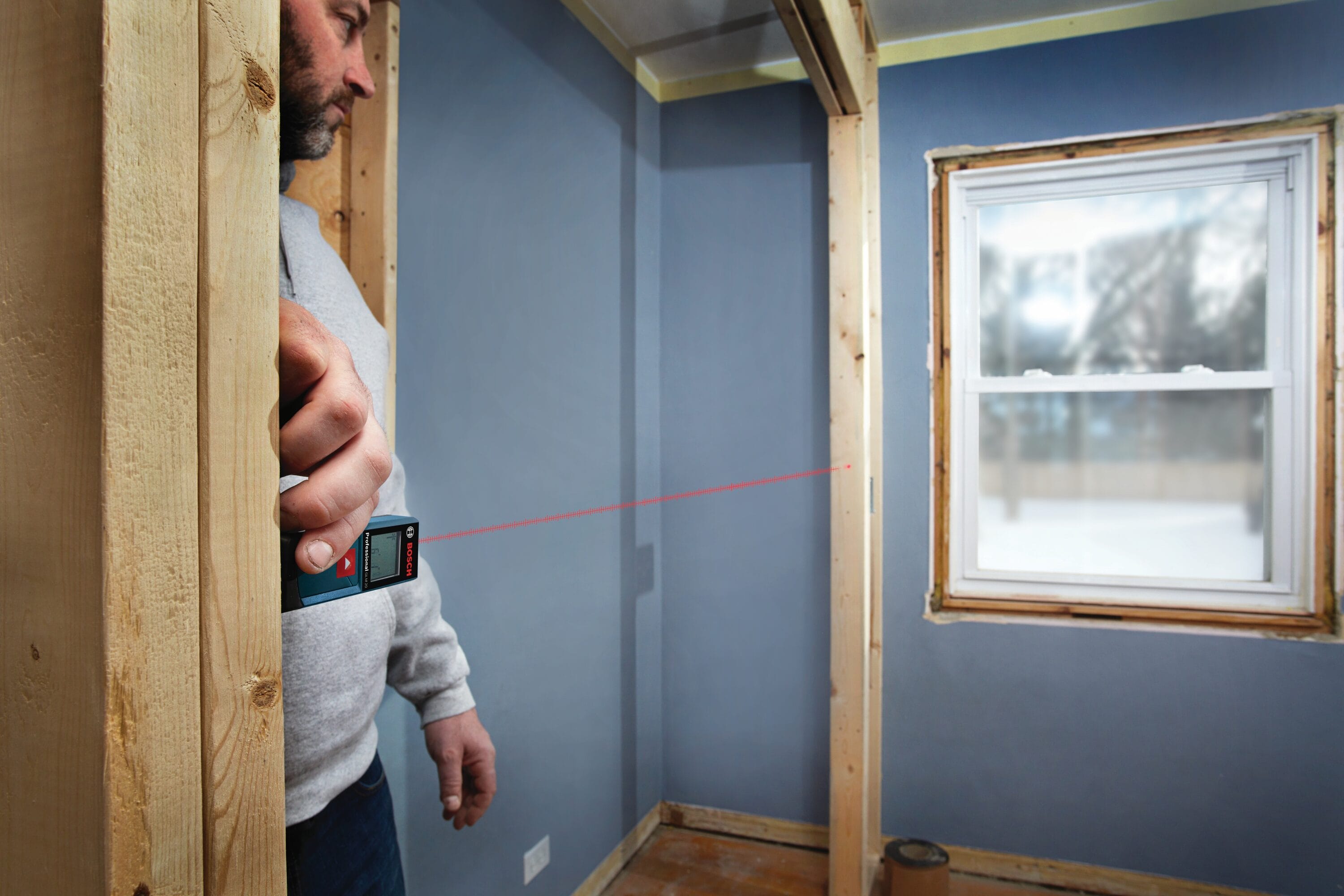 An application photograph of a residential carpenter holding the Bosch BLAZE laser measure tool flat against a structural wood wall stud to instantly verify horizontal dimensions across a room toward an exposed window framing gap.