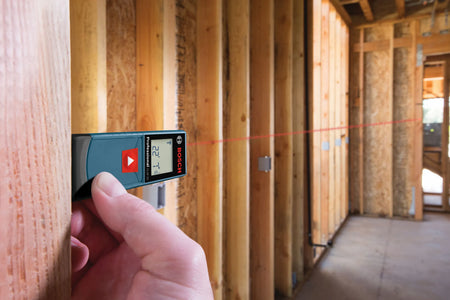 A close-up application shot focusing on a worker's hand gripping the Bosch GLM 20 laser measure tool horizontally against a vertical structural timber beam to project a red laser range alignment line down a long, open framing hallway.