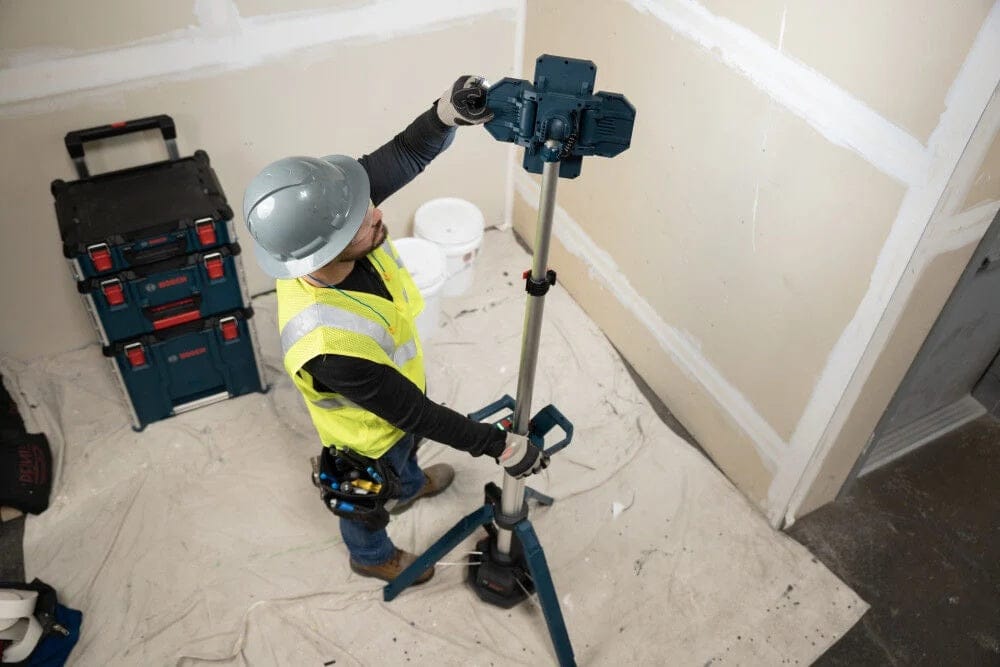 An overhead action shot of a worker in a yellow safety vest and hard hat adjusting the tilt of the LED light head on the fully extended tower light at a construction site.