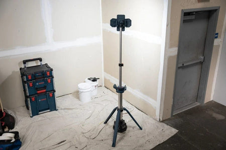 A wide shot of the Bosch tower light standing in a corner of an indoor workspace, providing powerful illumination for a job site with construction equipment visible in the background.