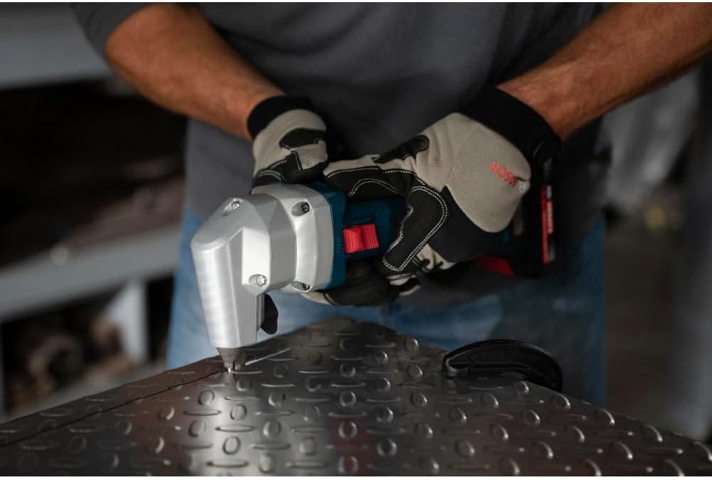 A close-up jobsite view of a metal fabricator wearing protective gloves firmly holding the Bosch brushless cordless nibbler horizontally to execute a clean, distortion-free line cut down a heavy sheet of steel diamond tread plate resting on a workbench.