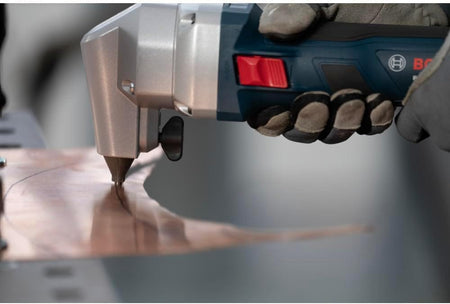 An operational action view of the Bosch brushless sheet metal nibbler being steered by a worker in safety gloves to punch a smooth, tight radiused scroll curve path out of an extended sheet of copper or thin brass paneling.