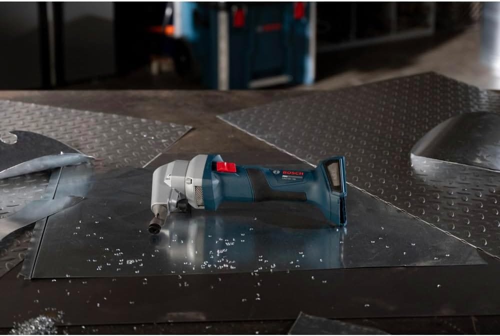 A wide jobsite workshop overview showing the compact Bosch cordless sheet metal nibbler resting flat on its side across a large sheet of reflective galvanized steel, surrounded by small crescent-shaped metal punch chips scattered on a workshop bench surface.