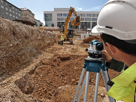 An outdoor jobsite application action photo showcasing a surveyor wearing a white hard hat and high-visibility vest as he peers through the eyepiece of the Bosch 26x optical transit level on its tripod, leveling out a deep dirt excavation pit where a coworker holds a measuring grade rod in front of a yellow hydraulic tracked excavator.
