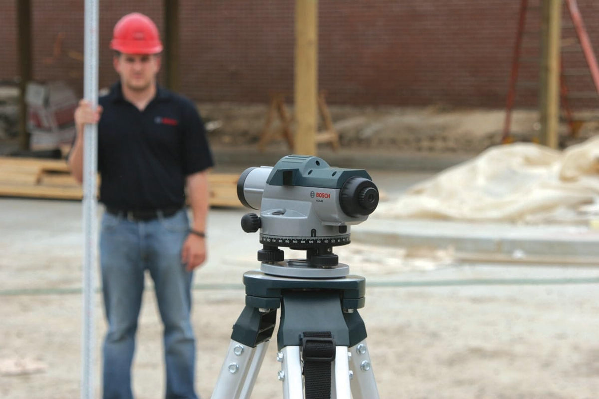 An outdoor application photo focusing on the back profile of the Bosch GOL26CK level tool mounted on its aluminum tripod, with a blurred surveyor wearing a red hard hat holding up a vertical grade rod in the background.